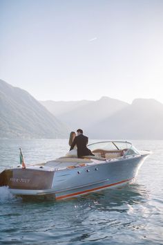 a man and woman riding on the back of a silver boat in water with mountains in the background