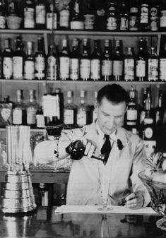 a man in a white suit pouring wine into a glass at a bar with liquor bottles behind him