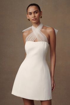 a woman wearing a white dress with sheer neckline and high low cut skirt, standing in front of a beige background