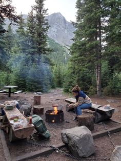 a woman sitting at a campfire in the middle of a forest filled with trees