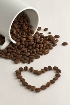 coffee beans spilling out of a white cup into the shape of a heart on a white surface