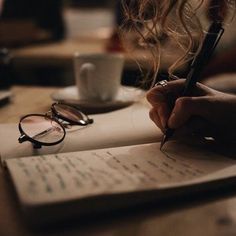 a person writing on a piece of paper next to a cup of coffee and eyeglasses