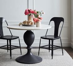 a white table with two black chairs and a vase filled with pink flowers on top