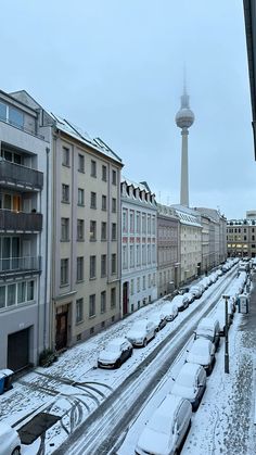 cars are parked on the side of a snowy road in front of tall buildings and a television tower