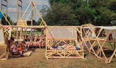 people sitting and standing around wooden structures on the ground with tents in the backgroud