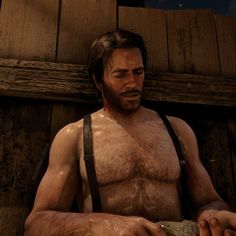 a man with no shirt and suspenders holding something in his hand while standing next to a wooden wall