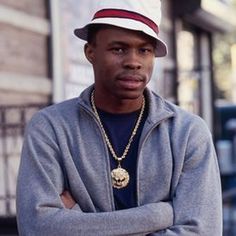 a young man with his arms crossed standing in front of a building wearing a hat