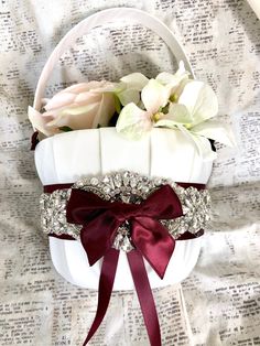 a white basket with red ribbon and flowers in it on top of a tablecloth