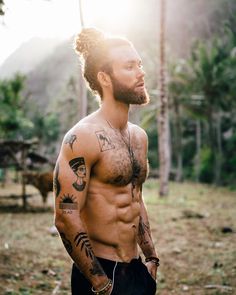 a shirtless man with tattoos on his chest standing in front of some trees and looking off into the distance