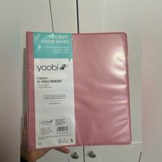 a person holding up a pink binder in front of a white wall with the words yoobi on it