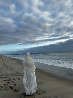 a woman walking on the beach with her head covered in a white shawl and looking out at the ocean