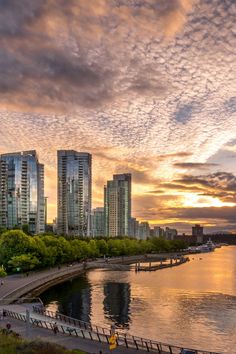 the sun is setting behind some tall buildings on the water's edge with people walking along it