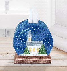 a cross stitch tissue holder with a house in the snow on it and a white towel next to it