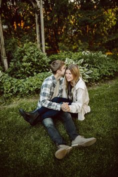 a man and woman sitting on the grass in front of some bushes with trees behind them