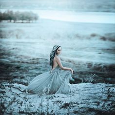 a woman in a long dress sitting on the beach looking at the water with her back to the camera