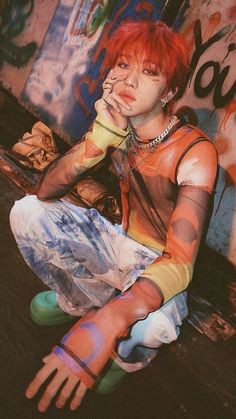 a man with red hair sitting on the ground in front of graffiti covered walls and holding his hands to his mouth