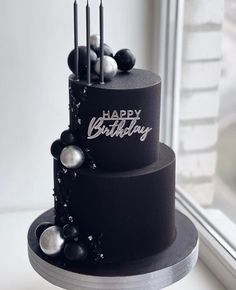 a black birthday cake with candles and decorations on the top is sitting in front of a window