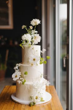 a three tiered cake with white flowers and greenery on the top is sitting on a wooden table