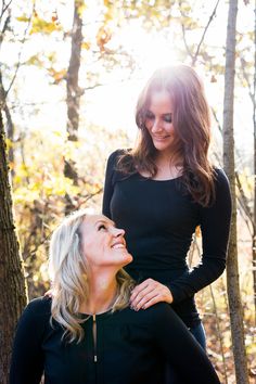 two women standing in the woods with their faces close to each other, smiling and looking at each other