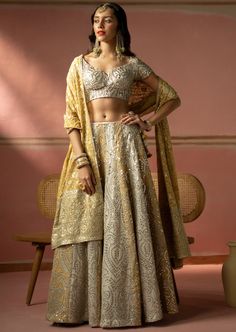 This lehenga features heavy silver zari embroidery with mirrorwork panels alternating between ecru and golden yellow color. The lehenga set shows delicate tassels detail. The short sleeves blouse has plunging neckline. It is paired with a mirror embroidered organza dupatta with tassels. Silver Lehenga With Mirror Work In Traditional Drape, Silver Sharara With Intricate Embroidery For Festivals, Silver Sharara With Intricate Embroidery For Diwali, Traditional Drape Silver Lehenga With Mirror Work, Traditional Silver Lehenga With Mirror Work, Silver Embroidered Sharara For Diwali, Silver Choli With Intricate Embroidery For Diwali, Silver Sharara With Mirror Work For Diwali, Silver Choli With Mirror Work For Festivals