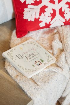 a book is laying on a blanket next to a red pillow and white snowflakes