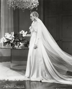 an old black and white photo of a woman in a wedding dress standing next to a table with flowers on it