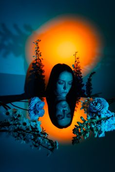 a woman's face is reflected in a mirror surrounded by flowers and trees with the sun behind her