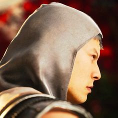 a close up of a person wearing a hood and looking off into the distance with red flowers in the background