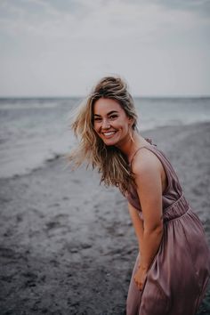 a woman standing on top of a sandy beach next to the ocean with her hair blowing in the wind