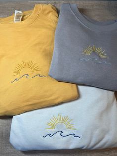 three t - shirts sitting on top of each other with the sun and waves embroidered on them