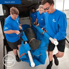 three men in blue shirts loading a large object into the back of a truck with another man looking on