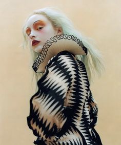 a woman with white hair and makeup wearing black and white clothing, standing in front of a beige wall