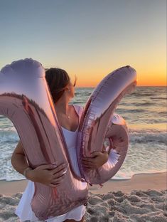 a woman on the beach holding two large balloons in front of her face as the sun sets