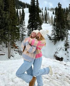 two women hugging each other in the snow with trees and mountains in the back ground