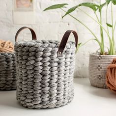 two woven baskets with leather handles sit on a white table next to potted plants