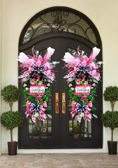 two large wreaths with pink flowers are on the front door of a house that is decorated with potted plants