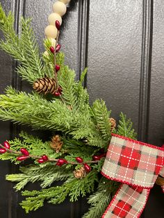 a christmas wreath with pine cones and plaid ribbon hanging from the front door on a black door