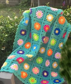 a crocheted blanket is sitting on a bench in front of some bushes and flowers