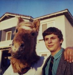 a man standing next to a horse mask in front of a house with a mans arm around him