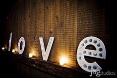 the word love is lit up in white letters on a wooden wall with candles around it