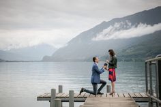 a man kneeling down next to a woman on a dock near the water with mountains in the background