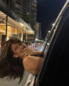 a woman leaning out the window of a car on a city street at night with buildings in the background