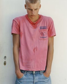 a young man standing in front of a white wall wearing a pink shirt and blue jeans