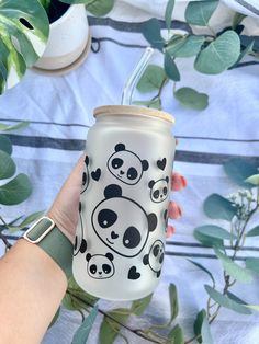 a hand holding a white and black cup with panda bears on it, surrounded by green leaves