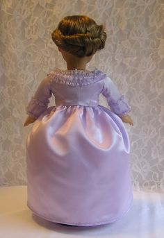 a doll in a purple dress sitting on top of a white tablecloth covered surface