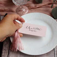 a person is holding a place card on a white plate with pink tassels