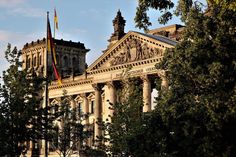 an old building with two flags on top and trees in the foreground against a blue sky