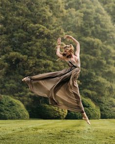 a woman in a long dress is dancing on the grass with trees in the background