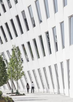 two people walking down the sidewalk in front of a large white building with many windows