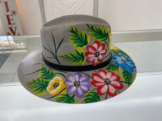 "Individually hand painted artisanal fedora hats. Each hat is meticulously hand painted by an artist. Each hat is unique and full of colors. Made by artists from Mexico the hats represent the beauty of Mexico.  A. Light gray with colorful flowers and leaf design. This hat is great for vacation in Tulum, Burning Man, Coachella, Beaches, or just brunching locally!  Made out of light canvas with air holes on top of hat for breathable and cool comfort. Size: Medium has elastic band on inside and can Handmade Artistic Fedora With Curved Brim, Handmade Multicolor Fedora For Spring, Artistic Hand-painted Fedora With Flat Brim, Artistic Fedora Sun Hat For Spring, Artistic Hand Painted Brimmed Panama Hat, Artistic Fedora With Short Brim For Spring, Handmade Artisan Panama Hat For Spring, Artisan Handmade Panama Hat For Spring, Handmade Multicolor Panama Hat For Spring